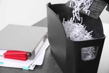 Image of a document shredder with paper shreds sitting on a table next to a closed binder and a stack of white papers.