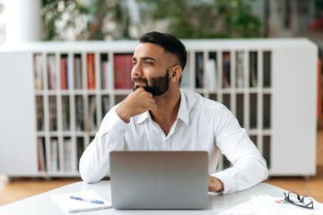 A businessperson who is sitting at a desk in an office is looking away and appears to be contemplating.