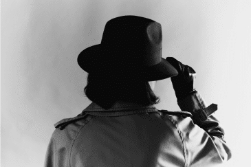 1940s-style black-and-white photo of the back of a female secret agent, from shoulders up, with trench coat, Fedora hat, and leather gloves