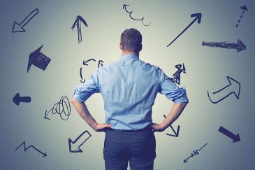 A confused business person stands with hands on hips while looking at many directional arrows drawn on a large whiteboard.