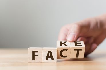 A person's hand is shown turning over wooden cubes with letters to change a spelled word from FAKE to FACT.