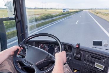 The driver is holding the steering wheel and is driving a truck on the highway. Empty road is ahead of him. The white truck is going in the opposite direction.